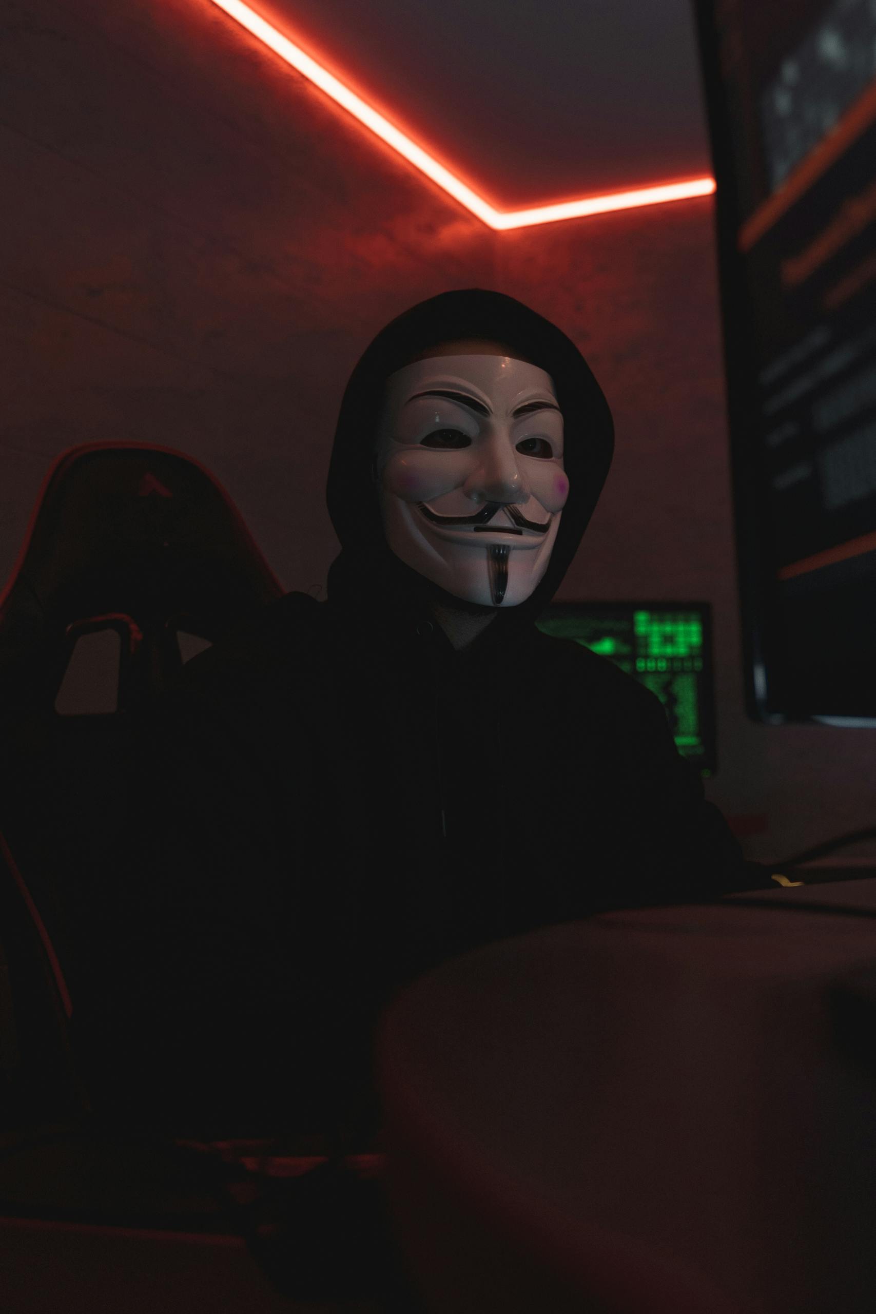 Mysterious figure wearing a Guy Fawkes mask, illuminated by computer screens in a dark room.