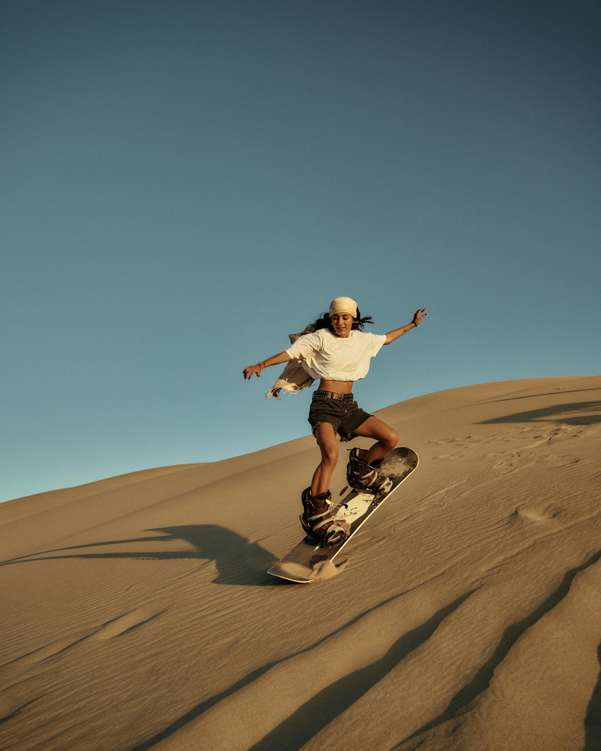 A young woman skillfully sandboards down a vast desert dune under a clear sky, showcasing adventure and thrill.