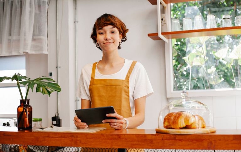 Sincere young female cafeteria worker with tablet looking at camera near container with tasty pastries